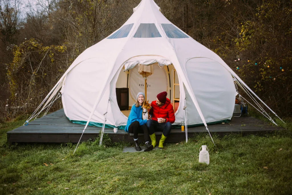 Couple of vacation goers campers sit in front of big glamping tent with their dog 