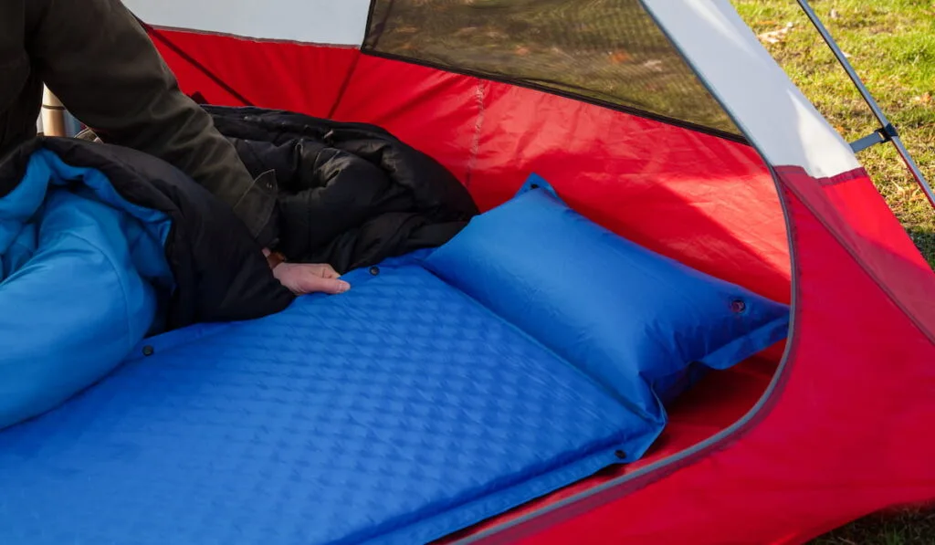A man gets his tent and sleeping bag ready at a campground by inflating and setting up his blue blow-up mattress pad to put for under his sleeping bag