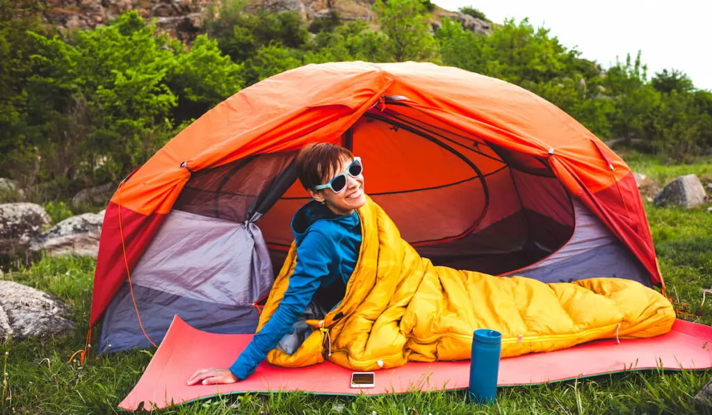 A woman sits near a tent in a sleeping bag against the background of the mountains
