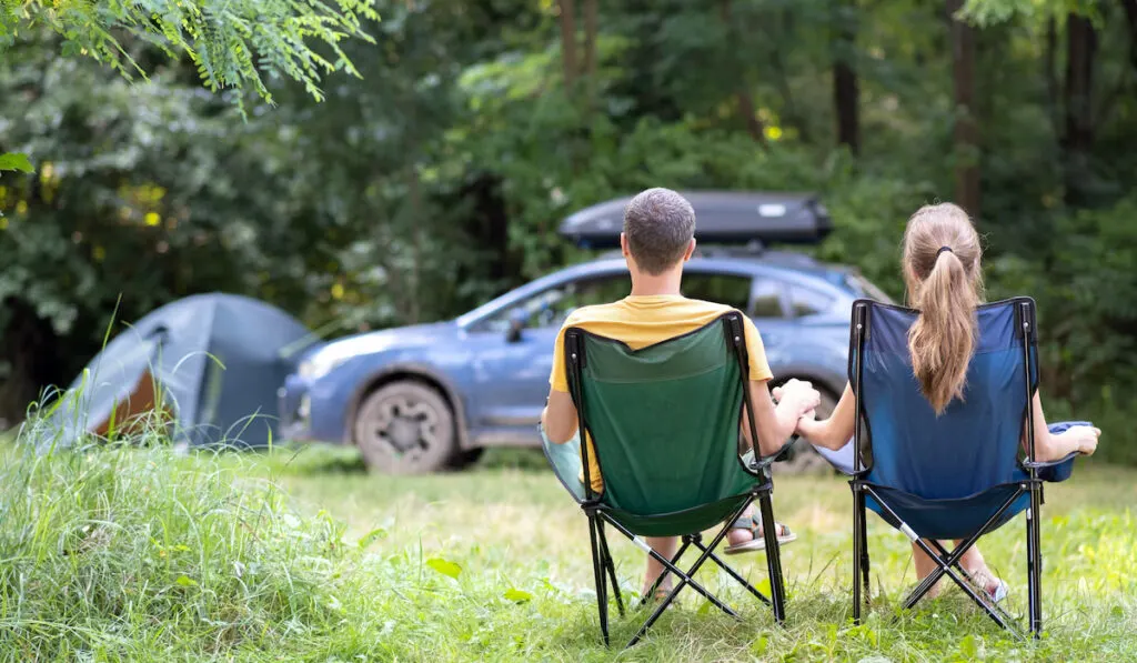 Back view of a happy couple sitting on camping chairs at campsite relaxing together.