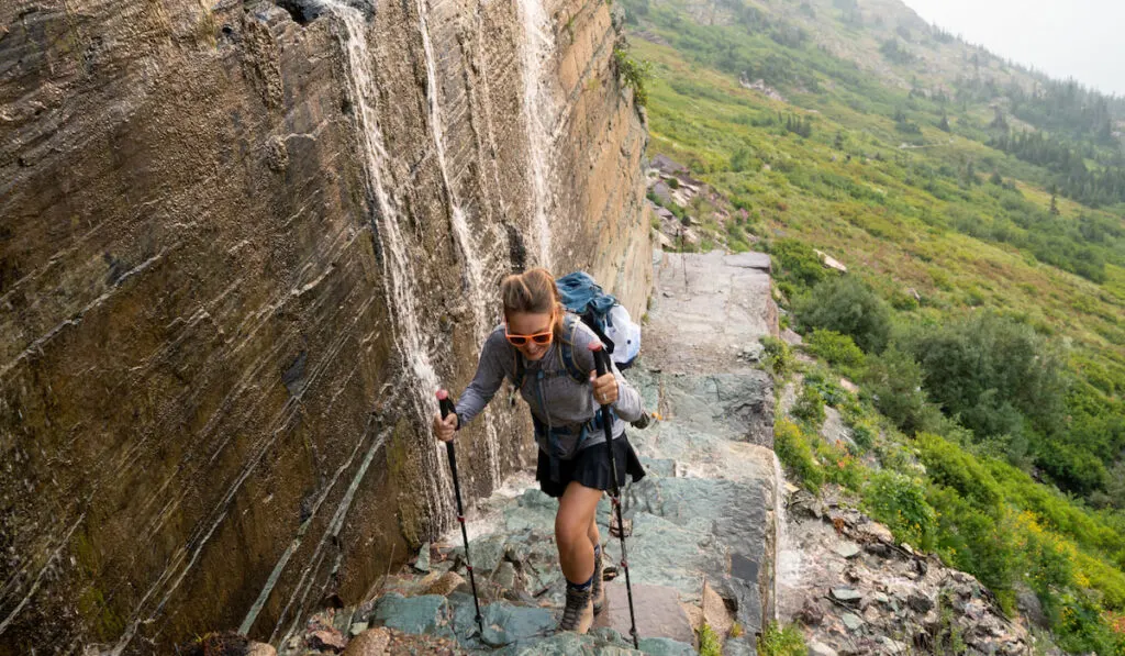 Brave woman hiker climbs up a narrow trail along a waterfall at the edge of a cliff 