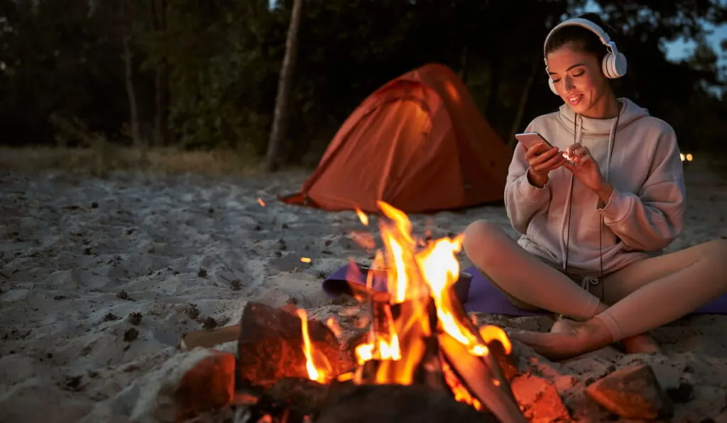 Cheerful young woman using cellphone while sitting neat campfire