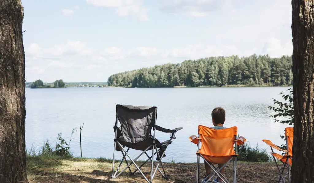 Child sitting in camping lounge chair on the riverbank in nature and looking at the view
