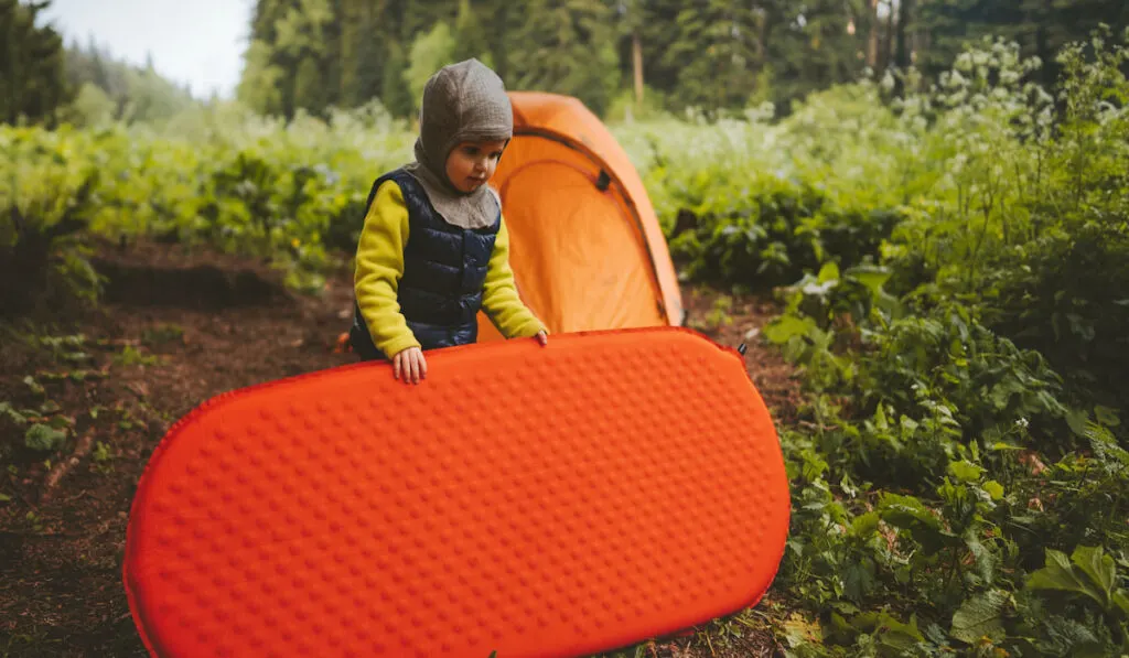 Child with sleeping pad helps to set camping tent 