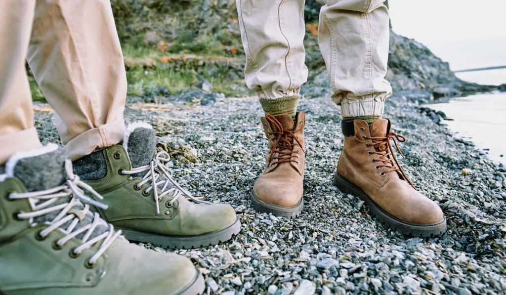 Close-up of unrecognizable couple standing in comfortable hiking boots on pebbles near water

