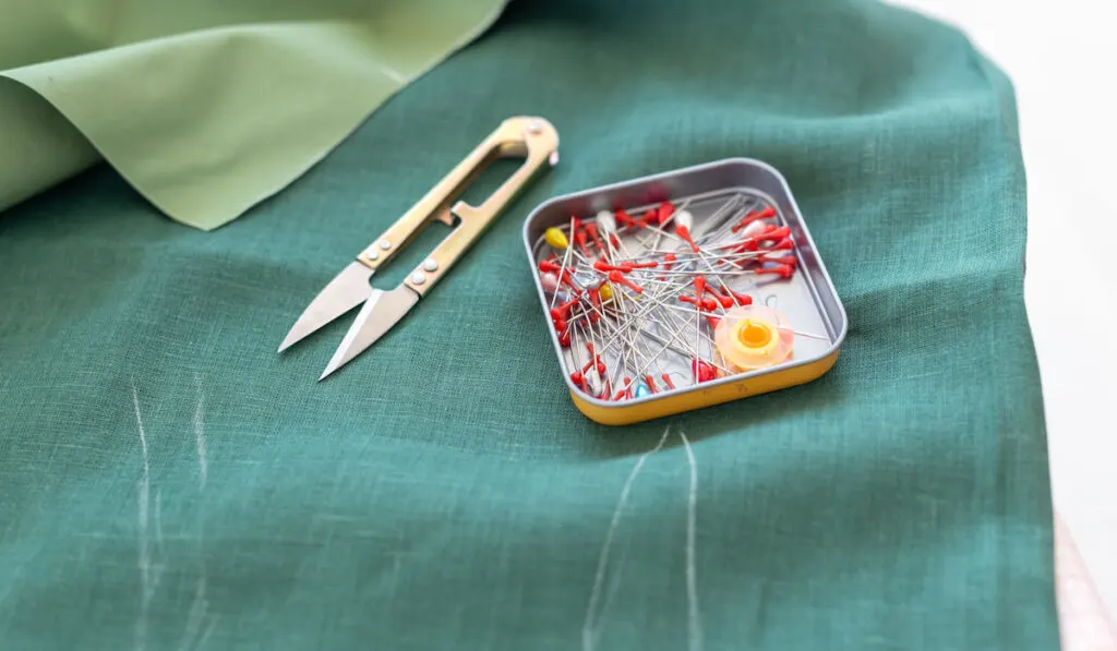 Close up safety pins and sewing scissors lie on a green piece of linen fabric