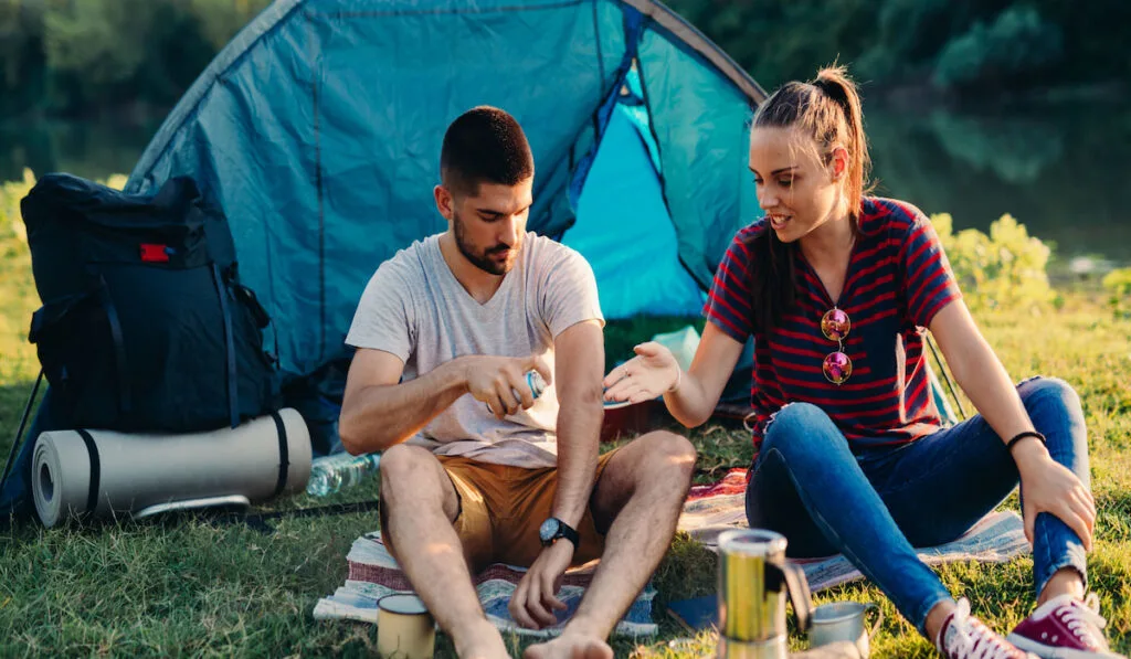 Couple camper spraying repellent for protecting from mosquito while camping near the river 