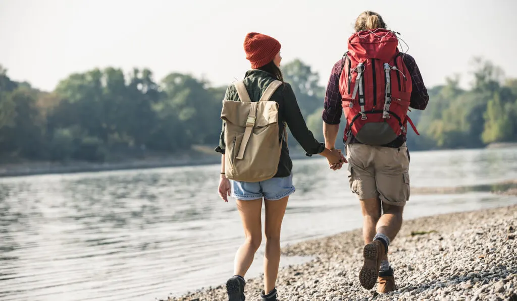 Couple hikers with backpacks walking hand in hand at the riverside
