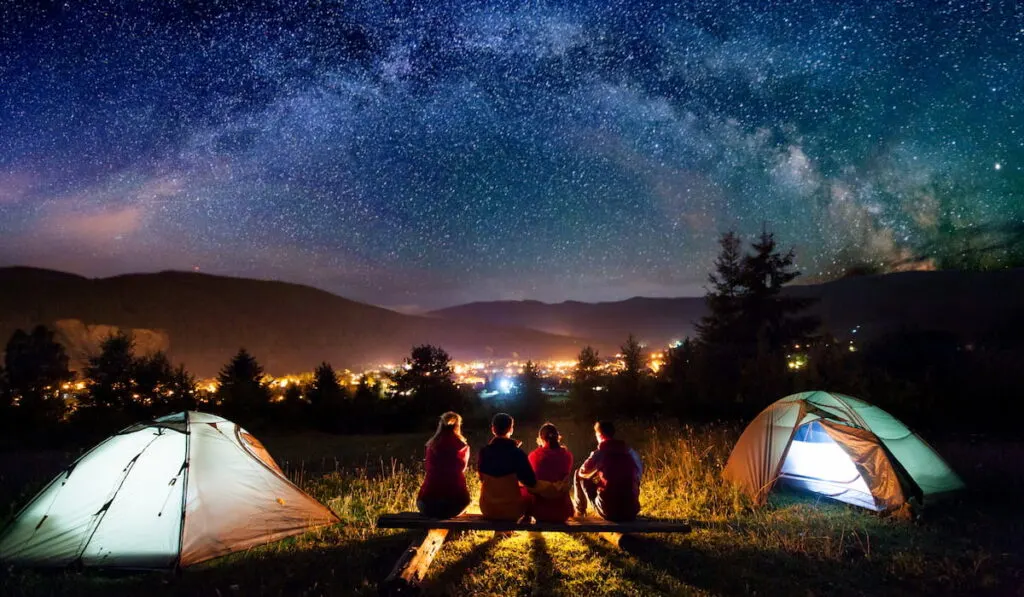 Friends hikers sitting on a bench made of logs and watching fire together beside camp and tents in the night