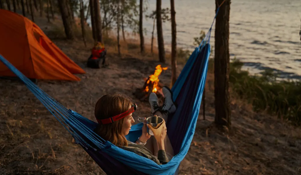 Hiker chilling out in hammock with hot tea at the end of day while camping in the woods beside lake