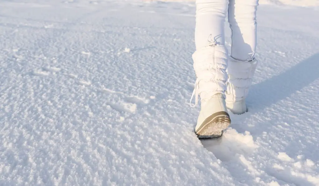 Low angle view of walking away female legs with white snow boots on, in deep snow.