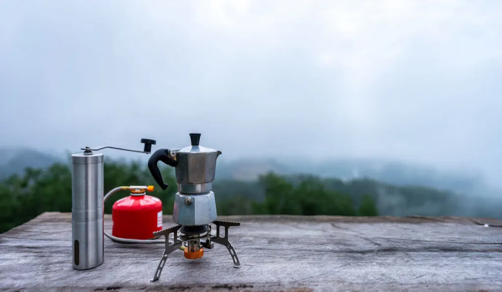 Portable coffee maker on fold-able gas stove and red gas tank with manual coffee grinder on wooden table in the middle of the forest