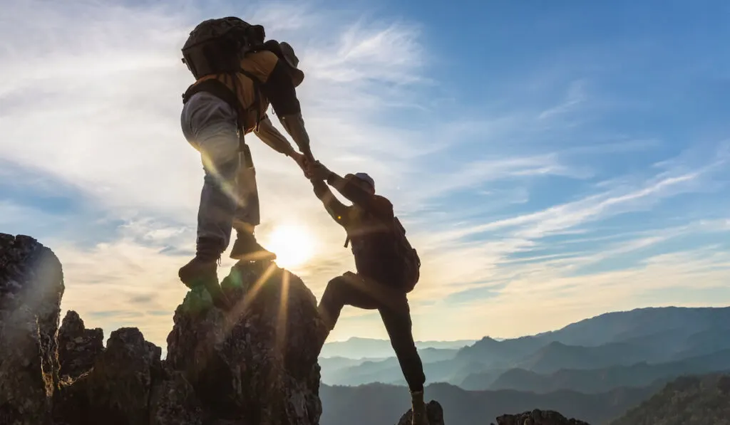 Silhouette Two Male hikers climbing up mountain cliff and one of them giving helping hand 