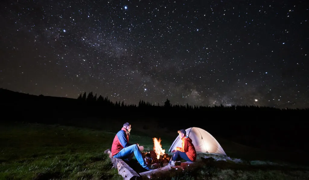 Tourists near campfire and tent under night starry sky
