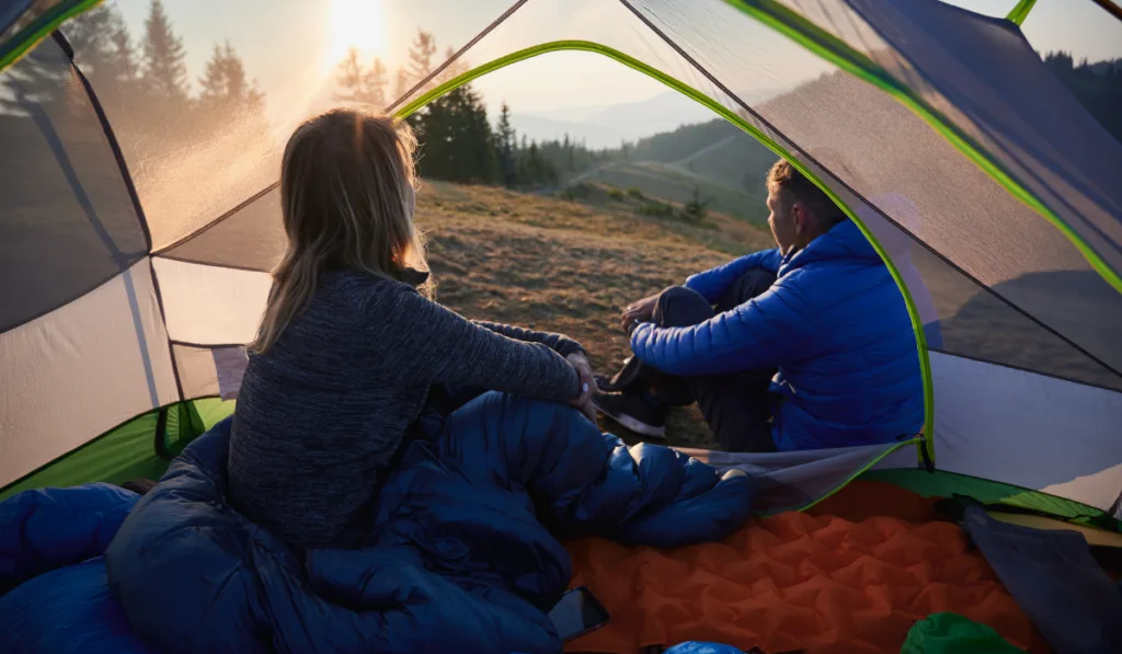 woman inside the camping tent and man outside the tent, both watching the sunset