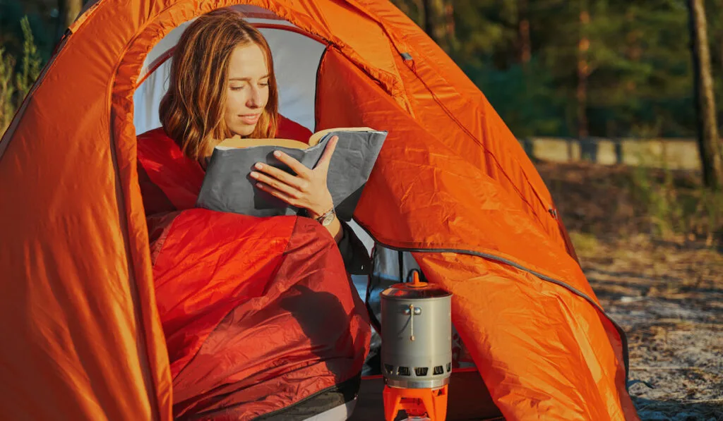 Woman camping, reading a book  while waiting for her tea prepared in kettle