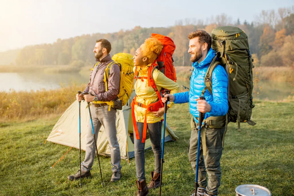 friends in colorful jackets hiking with backpacks, standing near the camping on the green lawn