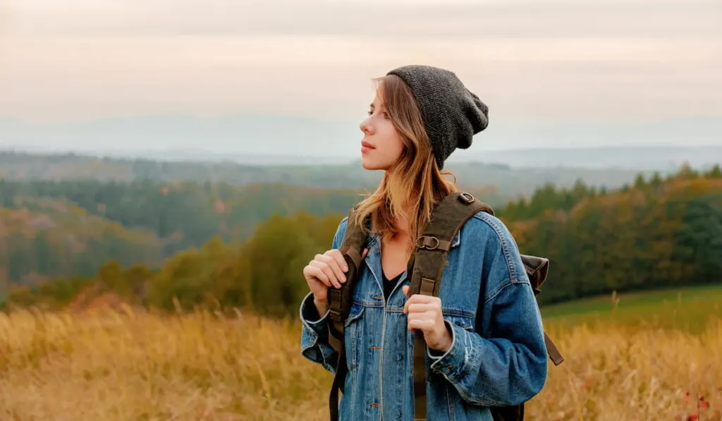 girl in denim jacket and hat with backpack in countryside with mountains on background 