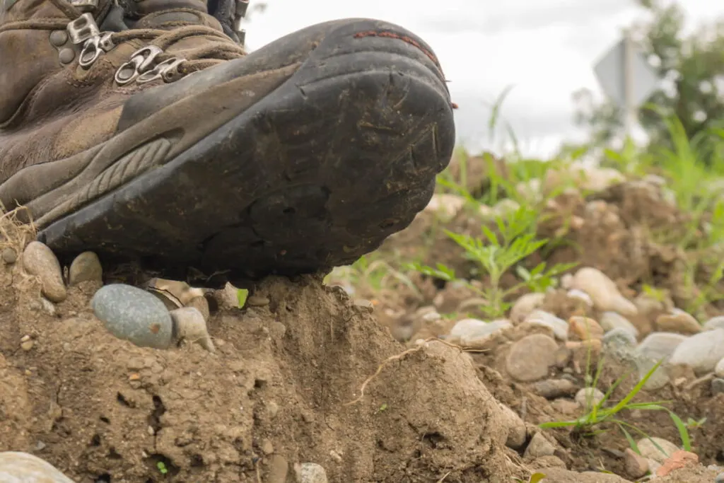 one (right) old worn hiking boot stands on a pile of gravel away from the road