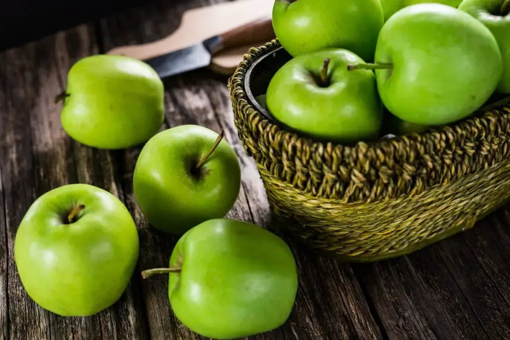 fresh green apples in a woven basket on top of a wooden table