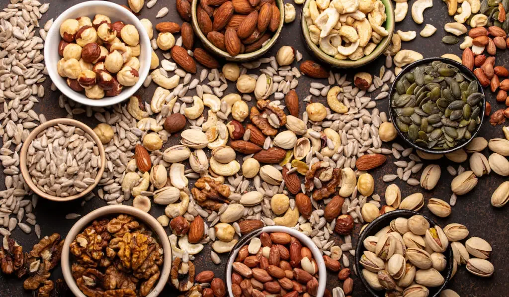 assorted raw nuts and various seeds in bowls on brown stone background