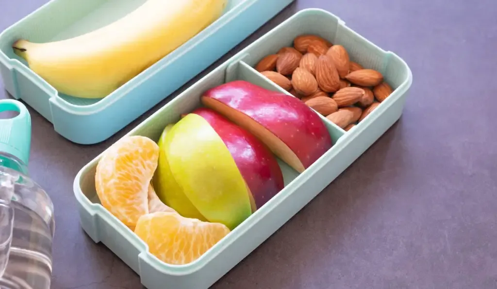 Fresh fruit in light green lunch box with almond, apple, orange, banana, water bottle on grey background.